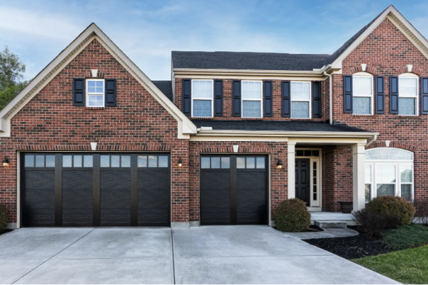 Two-story traditional brick home with black carriage-style double garage doors featuring upper window panels, white trim accents, dark shutters, and a wide concrete driveway.